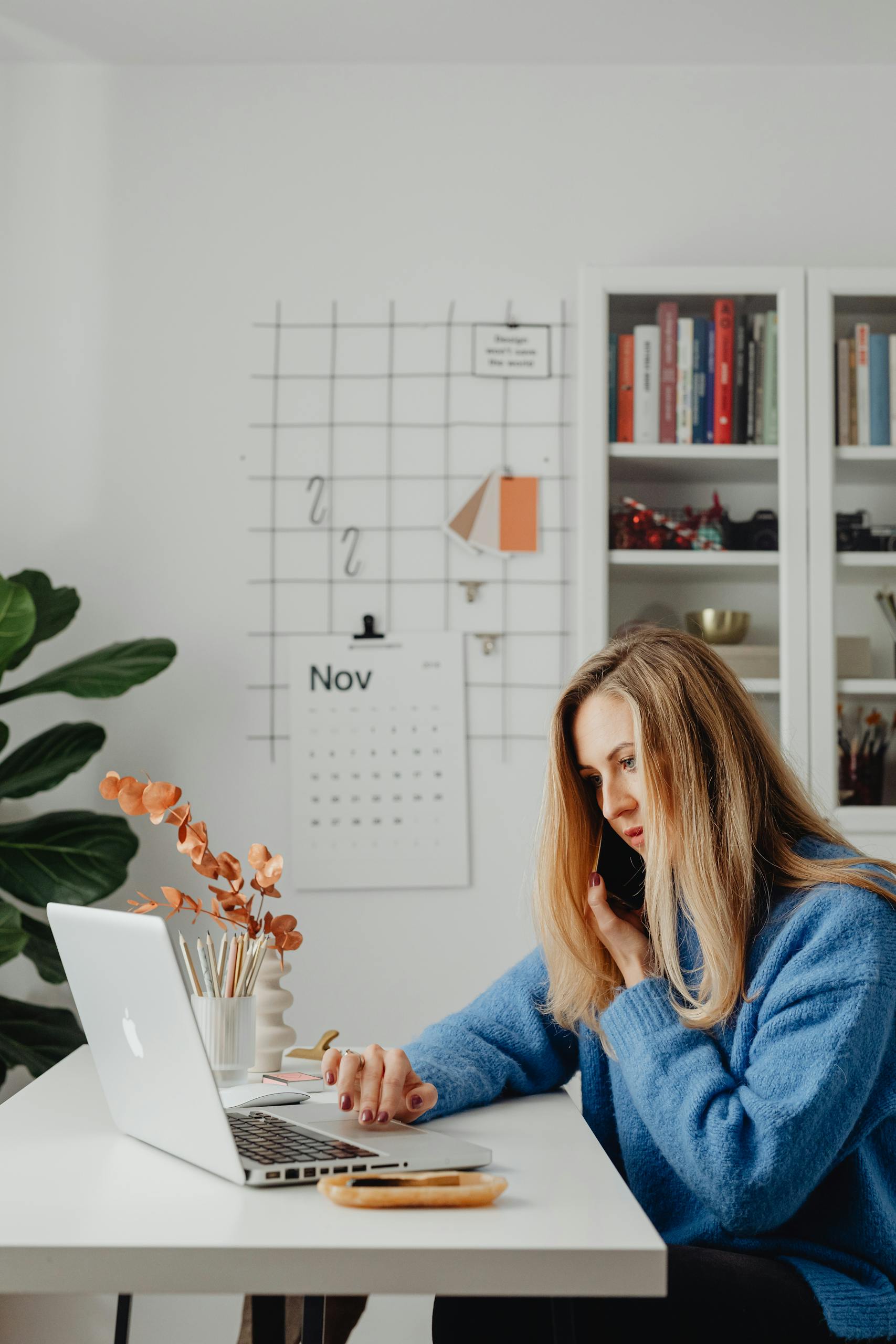 Woman multitasking at home office desk, using laptop and phone, surrounded by books and calendar.