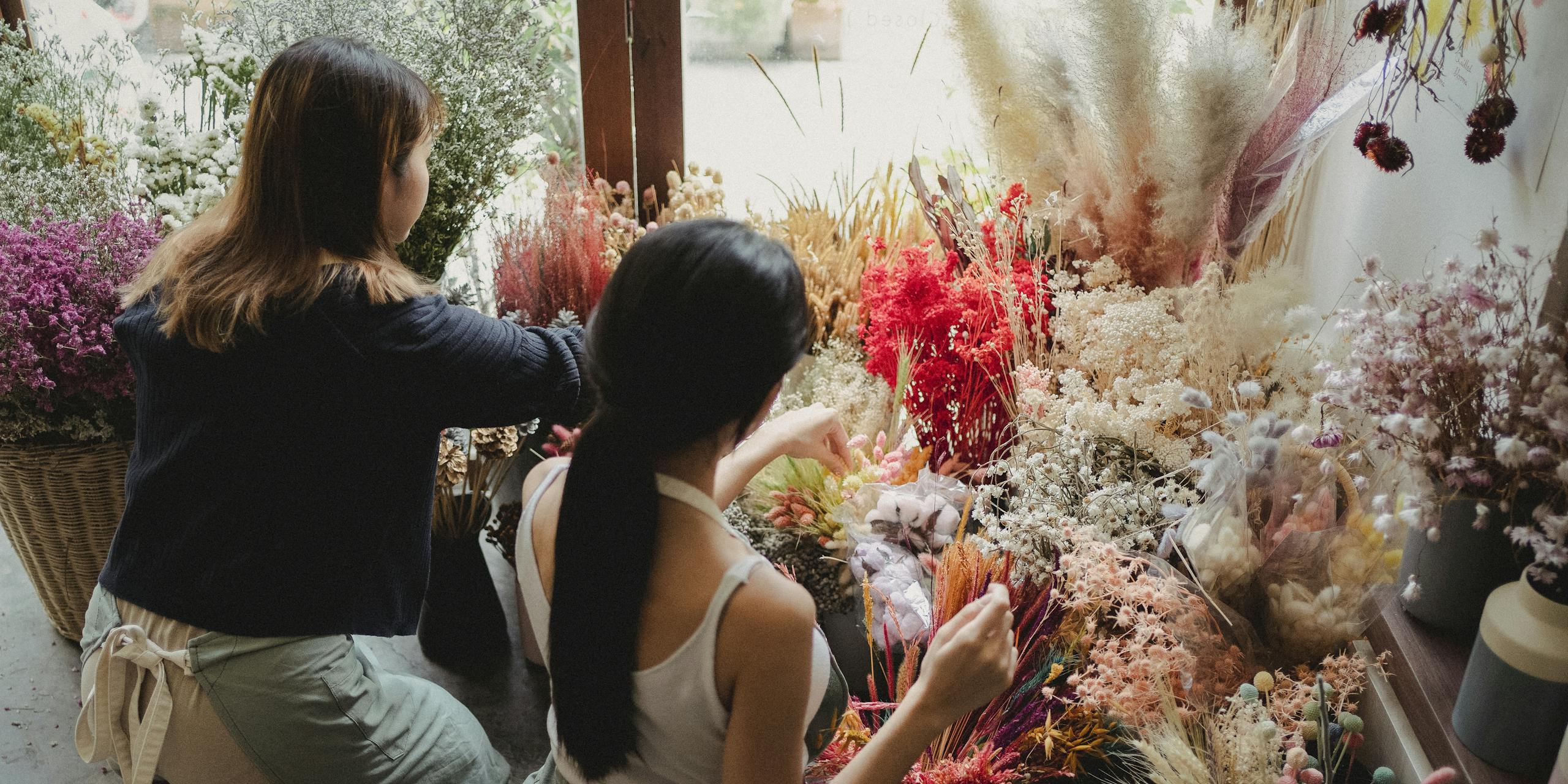 Back view unrecognizable female florists in uniforms sitting on floor near delicate flowers and arranging bouquets while working in florist shop